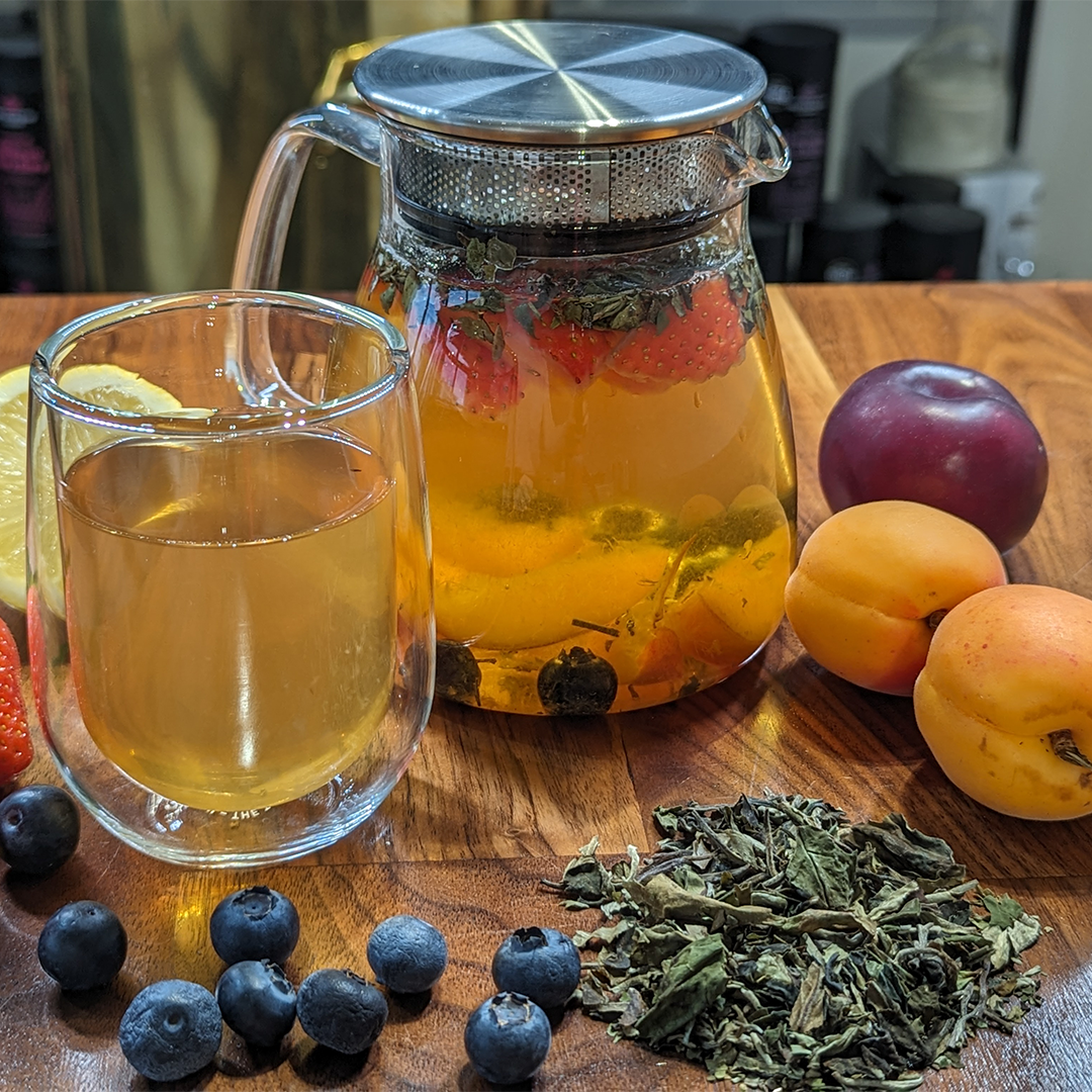 A glass teapot with infuser lid filled with cold brew tea and fruit. Surrounded by fruit, loose leaf tea and a full glass.
