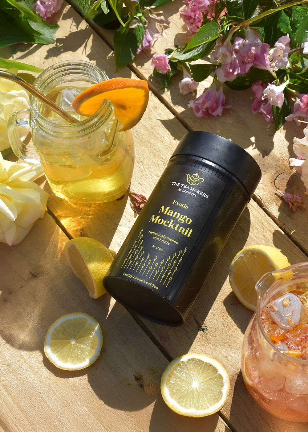 A black tin of fruit tea, surrounded by fruit tea in mugs, slices of lemon and flowers on a wooden picnic table.
