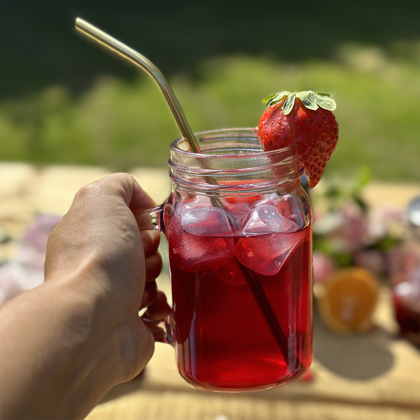 A hand holding an Fruity Iced Tea in a Mason Jar Mug with a Strawberry on the rim and a straw.