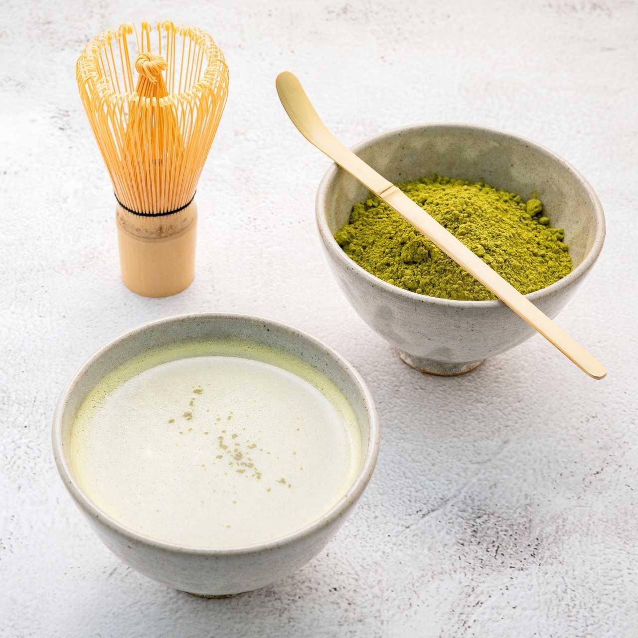 Bamboo whisk and spatula next to two bowls of matcha brewed and powder